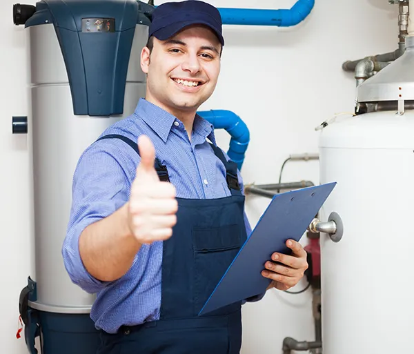 Smiling male plumber giving a thumbs-up gesture while holding a clipboard in a utility room with plumbing equipment.