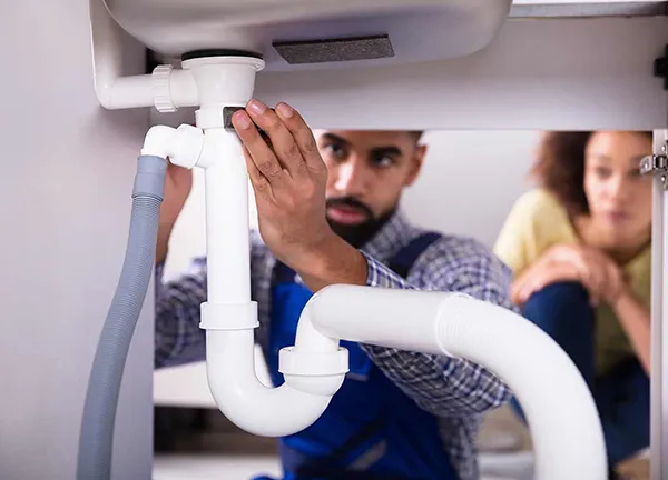 Plumber inspecting and repairing pipes under a kitchen sink while a customer watches attentively.
